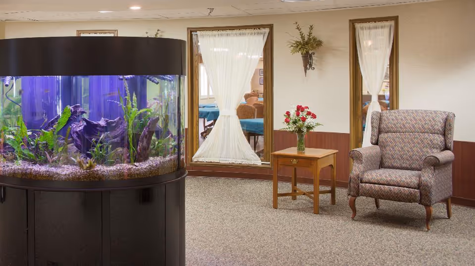 A cozy interior room featuring a large cylindrical aquarium filled with plants and fish on the left, a wooden side table with a vase of red and white flowers in the center, and a patterned upholstered armchair on the right. Behind the furniture are two large windows with sheer white curtains and a wall-mounted plant holder with greenery.