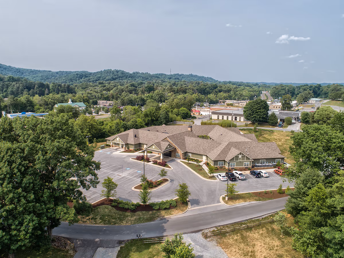 Aerial view of Dominion Senior Living of Anderson, showing a large single-story building with a brown roof surrounded by parking lots, trees, and greenery. The facility is situated in a semi-rural area with hills and other buildings in the background under a partly cloudy sky.
