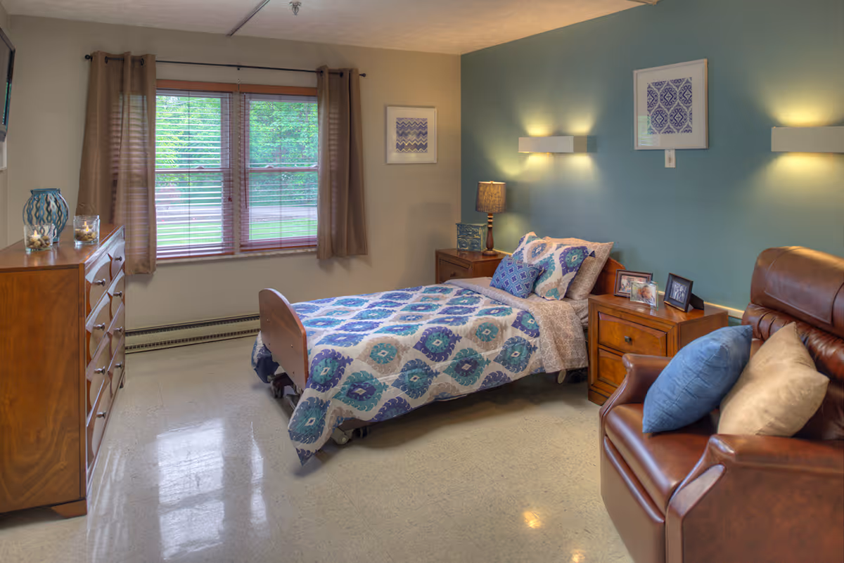 Furnished bedroom with a single bed covered in patterned bedding, wooden dresser and nightstands, a recliner chair, and a window with curtains.