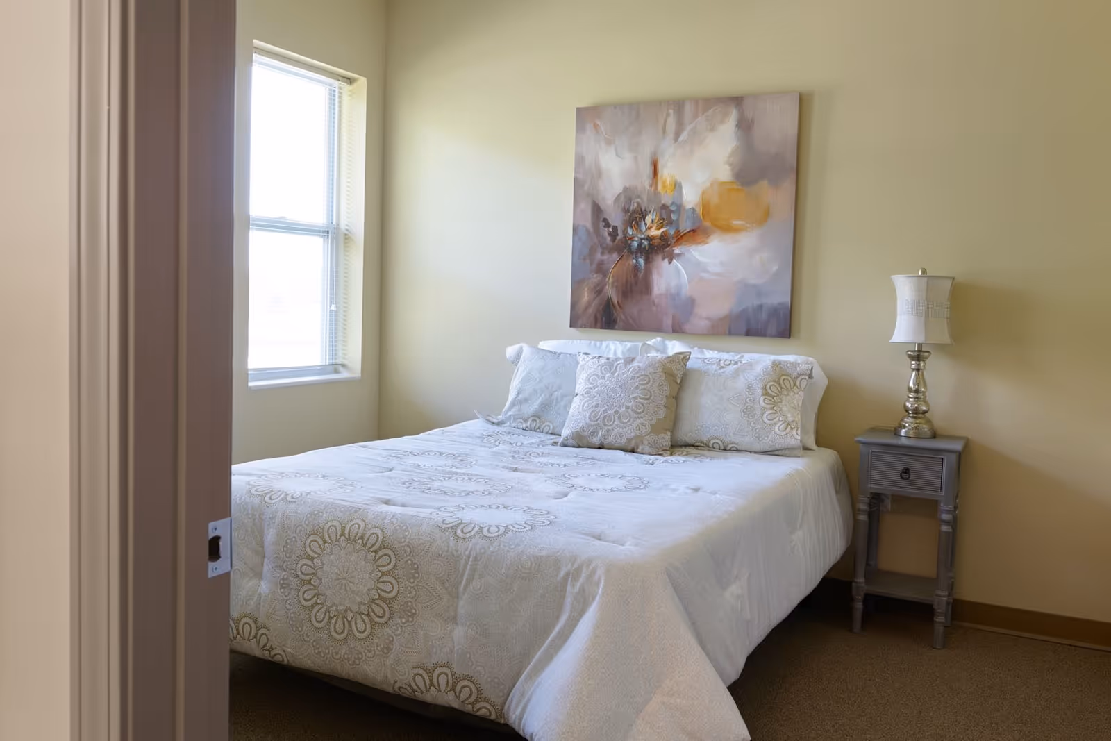 A bedroom with a neatly made bed featuring white and beige patterned bedding and pillows. There is a window with blinds on the left wall, a nightstand with a decorative lamp on the right side of the bed, and a large abstract painting hanging above the bed on a light yellow wall.