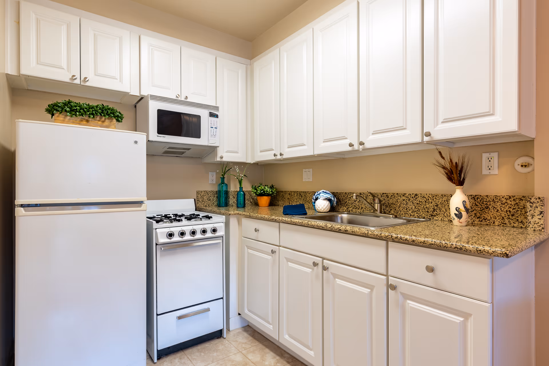 Compact kitchen with white cabinets, a refrigerator and stove, microwave, granite countertops, sink, and decorative plants.