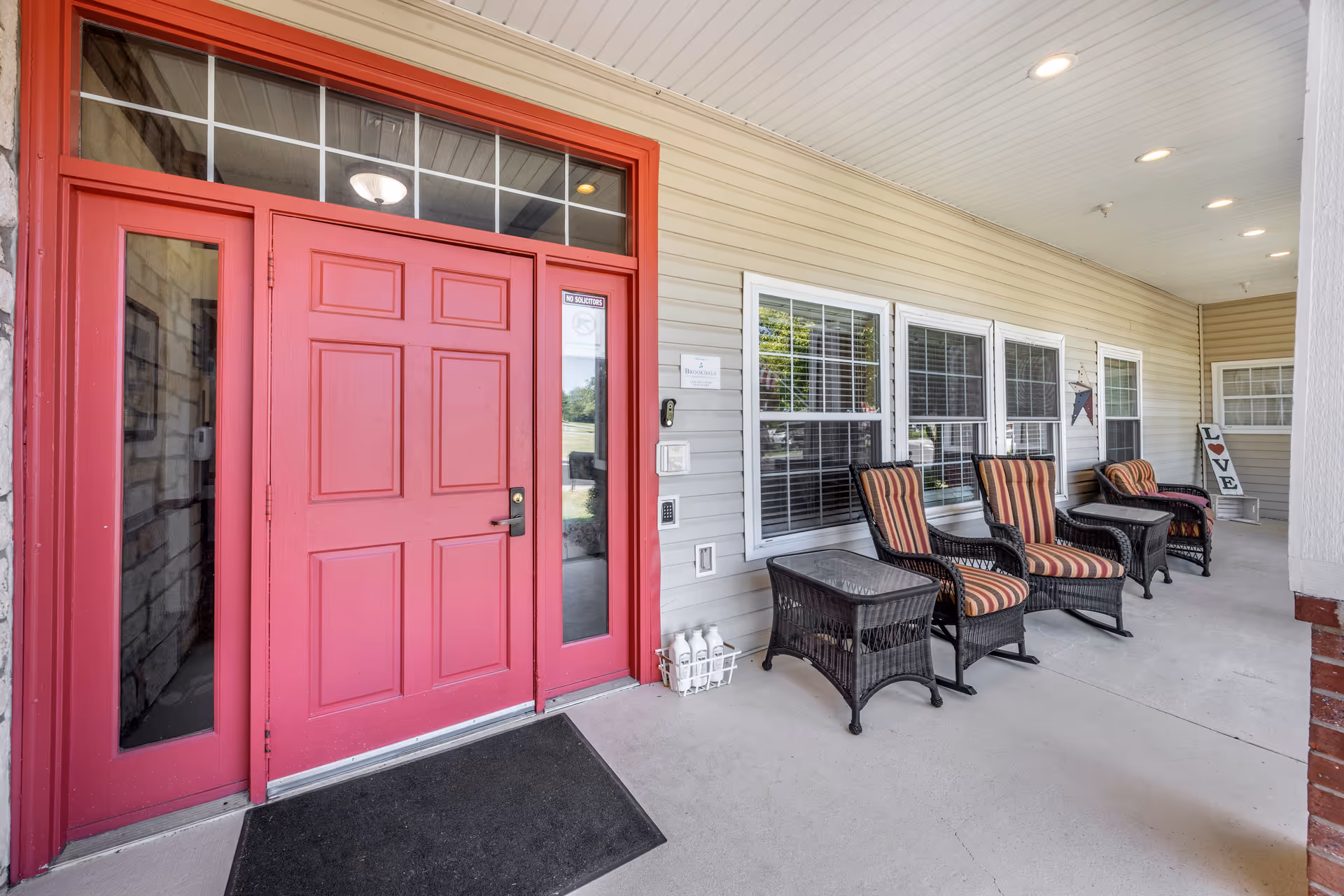 Covered porch area with a bright red front door and sidelights. Four wicker chairs with striped cushions and two wicker side tables are arranged along the wall with windows. A decorative sign spelling 'LOVE' is placed near the far end of the porch. The porch ceiling has recessed lighting.