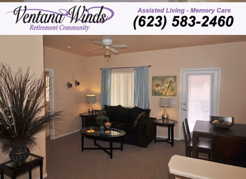 Interior view of a living room area in a retirement community featuring a black sofa with cushions, a round glass coffee table, a side table with a lamp, a dining table with chairs, a ceiling fan, and a window with curtains. There is also a potted plant on a small table and a door leading outside.