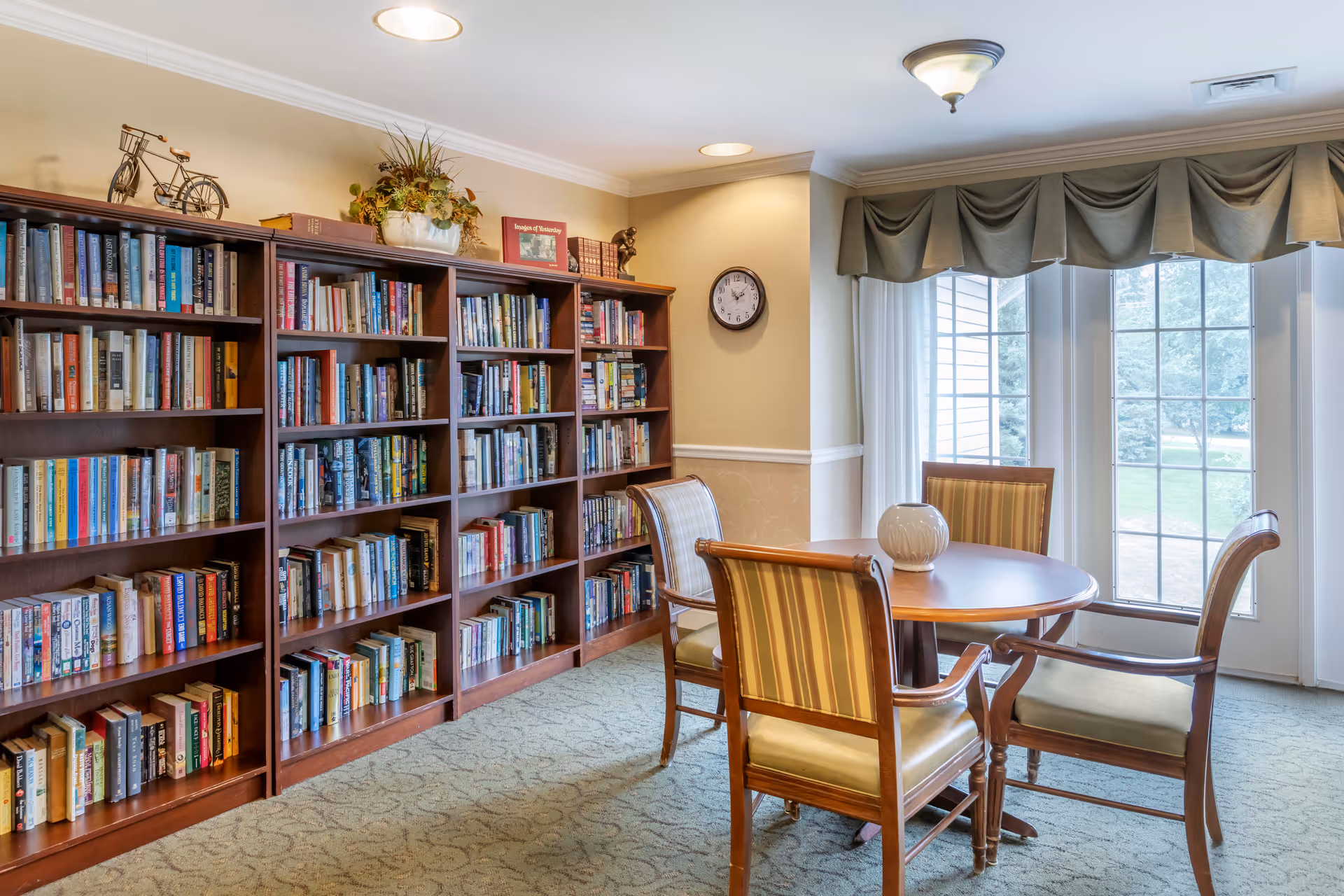 A cozy reading area in a senior living facility with a large wooden bookshelf filled with books, a round wooden table with four striped cushioned chairs, a decorative vase on the table, a wall clock, and large windows with draped curtains letting in natural light.