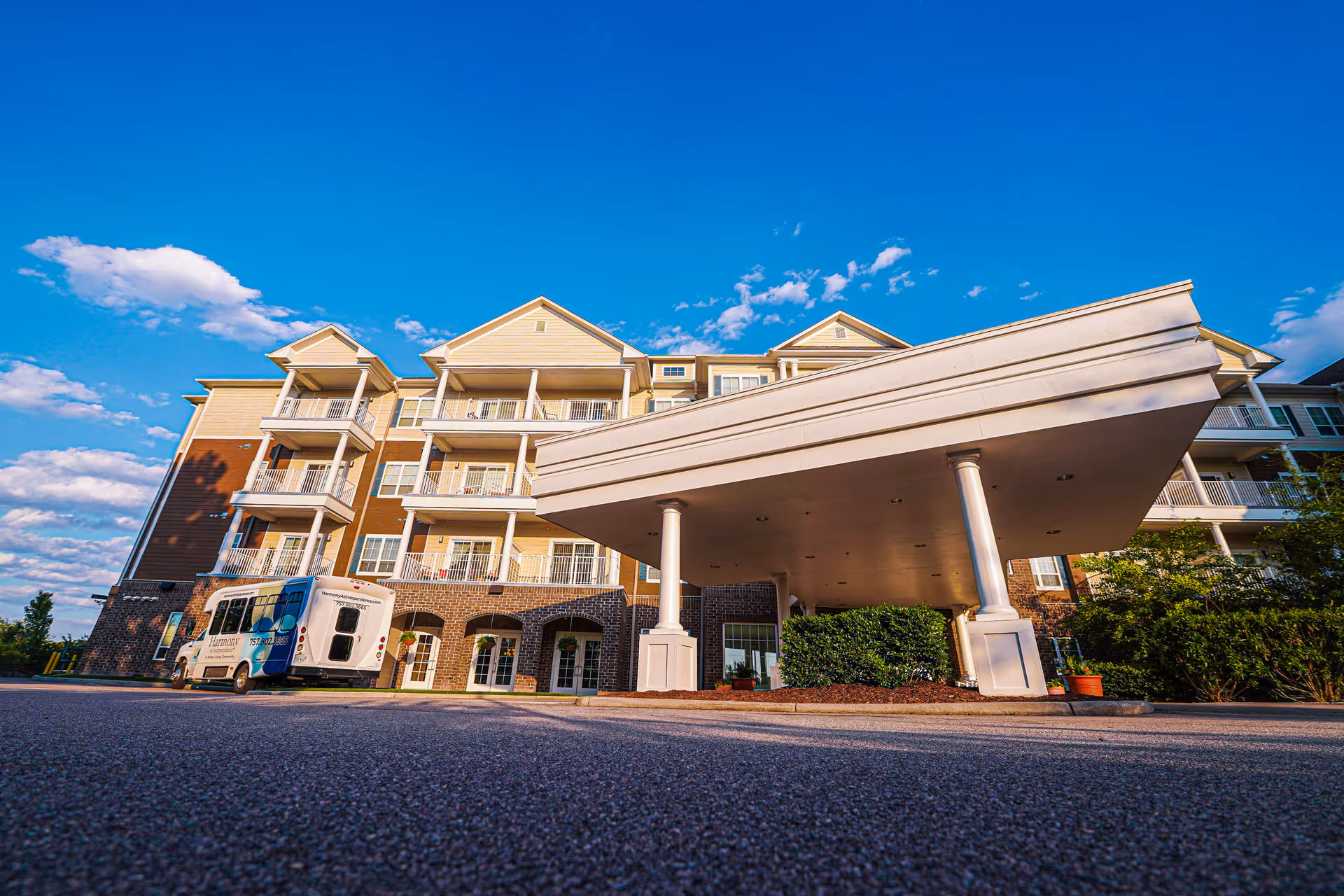 Entrance canopy and front facade of a multi-story senior living building with balconies and a parked shuttle van under a blue sky.