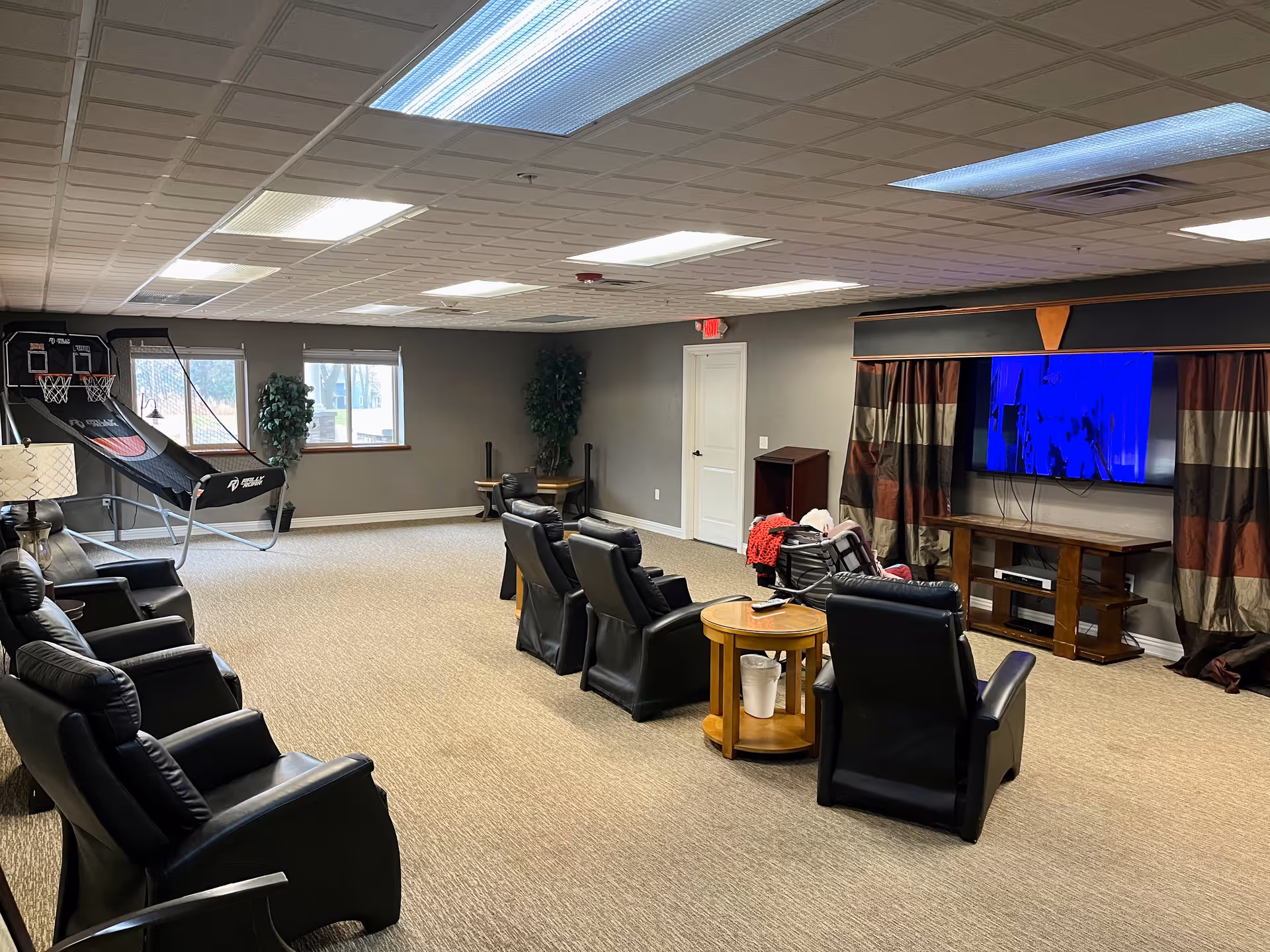 A spacious common room with several black leather recliners arranged facing a large flat-screen TV mounted on the wall. The room has beige carpet, gray walls, and a drop ceiling with fluorescent lighting. There are two windows with white blinds, two potted plants, a small wooden table with a lamp, and a basketball arcade game in the corner. A person is seated in one of the chairs near the TV.