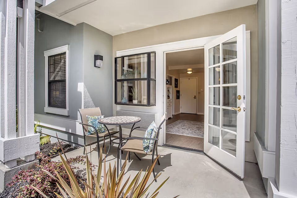 Outdoor patio area with a small round table and two chairs with patterned cushions. The patio is adjacent to a building with light gray walls and a white-framed window. A white-framed glass door is open, leading into an interior hallway with a rug and a closed door at the end.
