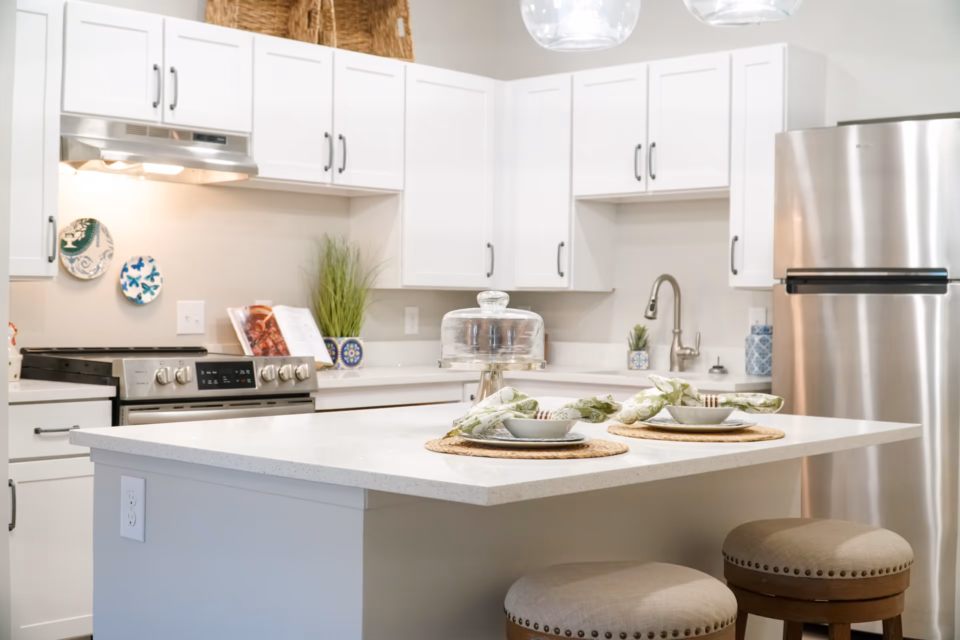 Bright modern kitchen with white cabinets, stainless steel refrigerator and stove, a white island countertop with two place settings, and two cushioned stools. Decorative plates and plants are placed on the countertops and walls.
