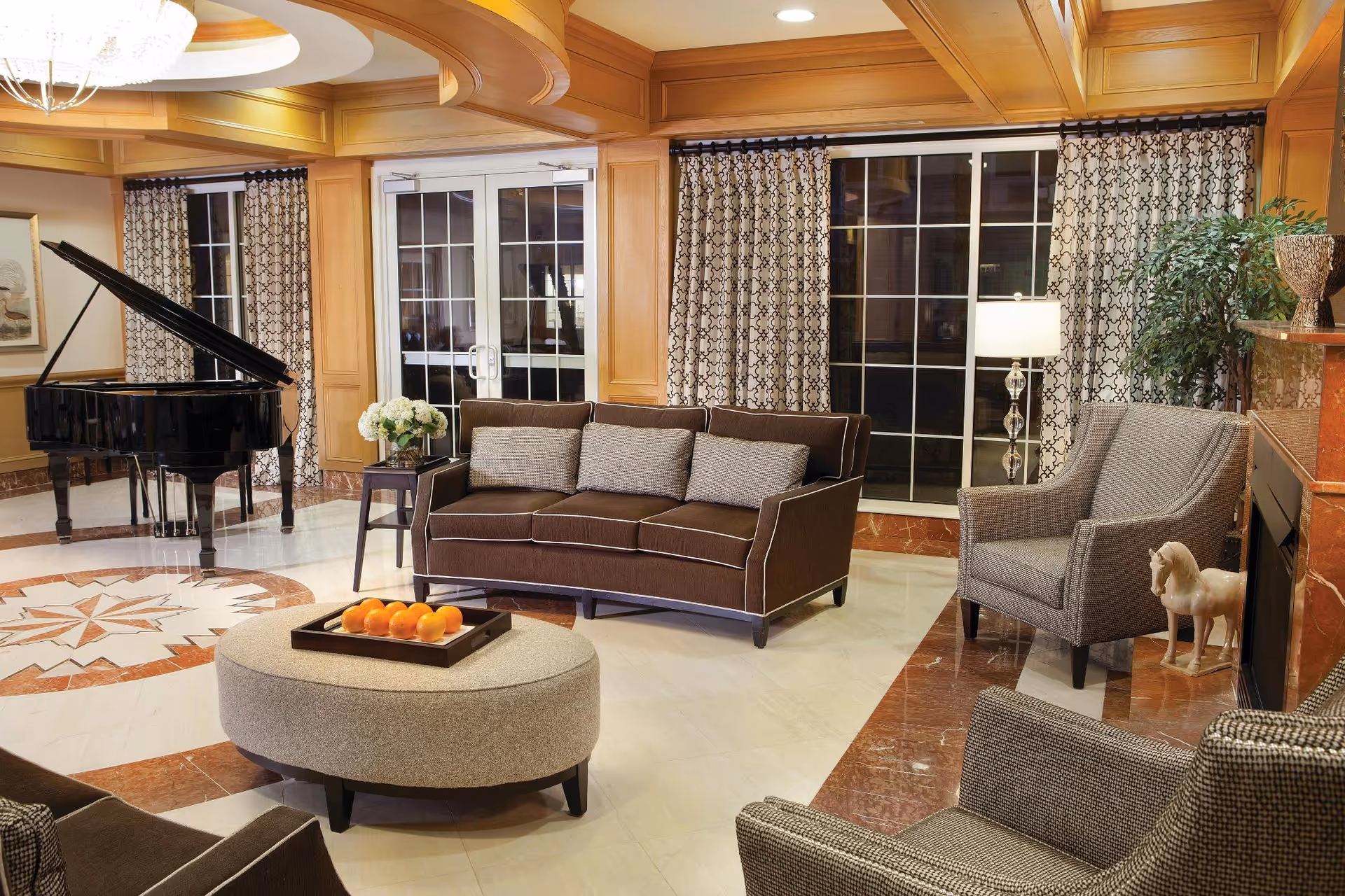 Elegant communal lounge with a sofa, armchairs, round ottoman, grand piano and large windows under a decorative ceiling.