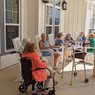 A group of elderly women sitting and chatting on white rocking chairs on a covered porch. One woman is in a wheelchair, and another has a walker nearby. The porch has beige siding and windows with black shutters.