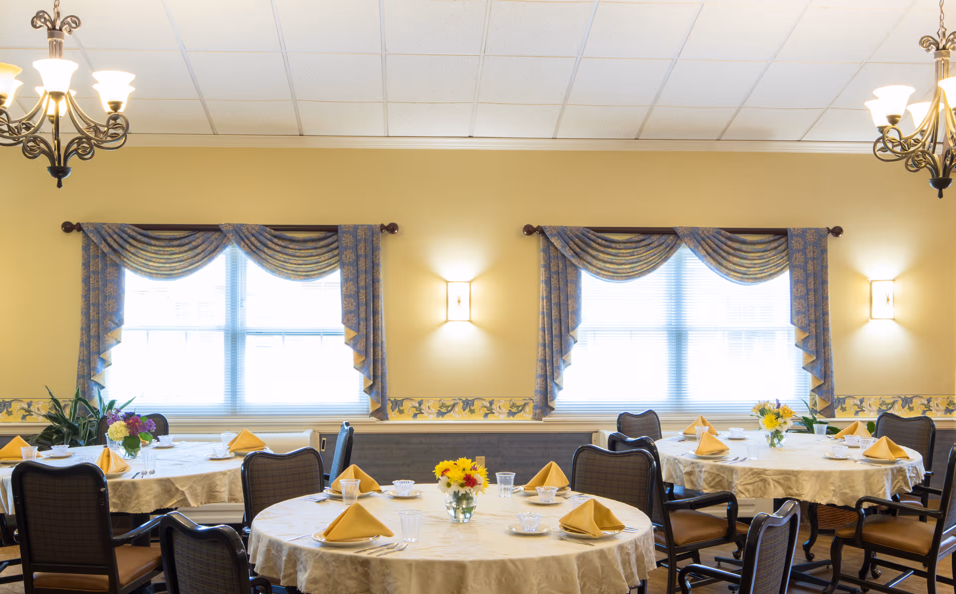 Bright dining room with round tables set with yellow napkins, floral centerpieces, chairs and draped windows.