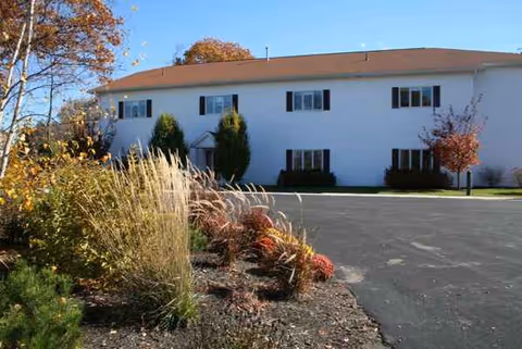 White two-story senior living facility with landscaped shrubs and a paved parking lot, front view.
