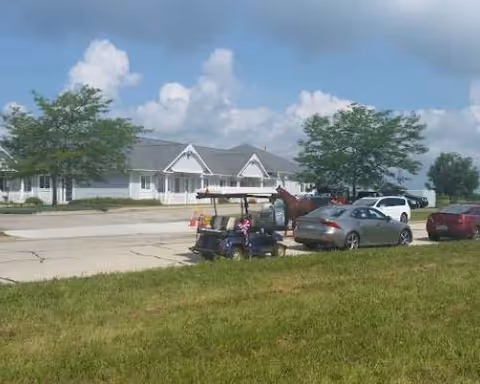 Single-story white residential building with pitched roofs, parked cars and a golf cart on the driveway and grass in the foreground under a blue sky.