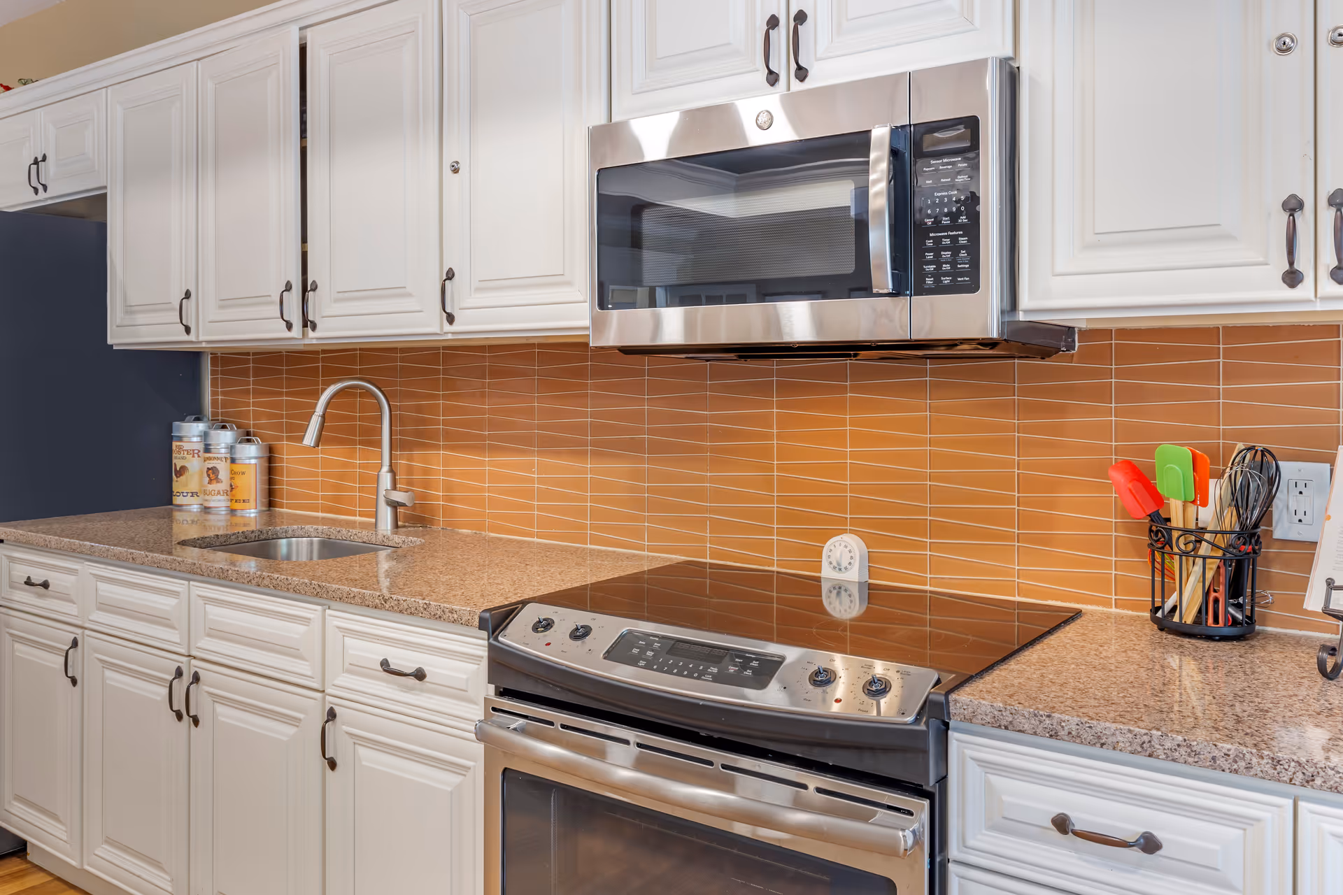 Modern kitchen with white cabinets, a stainless steel microwave above a black electric stove, a granite countertop with a sink and faucet, orange tiled backsplash, and kitchen utensils in a holder.