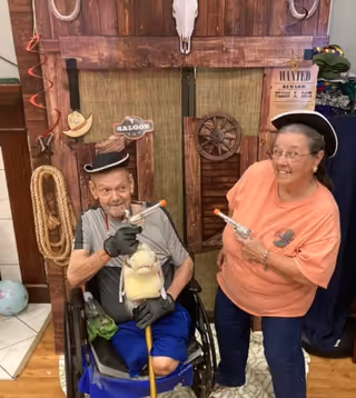 An elderly man in a wheelchair and an elderly woman standing next to him, both wearing hats and holding toy guns, posing playfully in front of a western-themed backdrop with a wooden door, a rope, a cowboy hat, a wagon wheel, and a 'Wanted' poster.