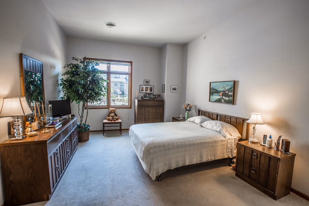 A cozy bedroom with a large bed covered in a white bedspread, flanked by two wooden nightstands each with a lamp. There is a wooden dresser with a mirror on the left side, a small TV, and decorative items. A large window lets in natural light, and a potted plant stands beside it. A chair with a teddy bear sits near the window, and framed pictures and a painting hang on the walls.