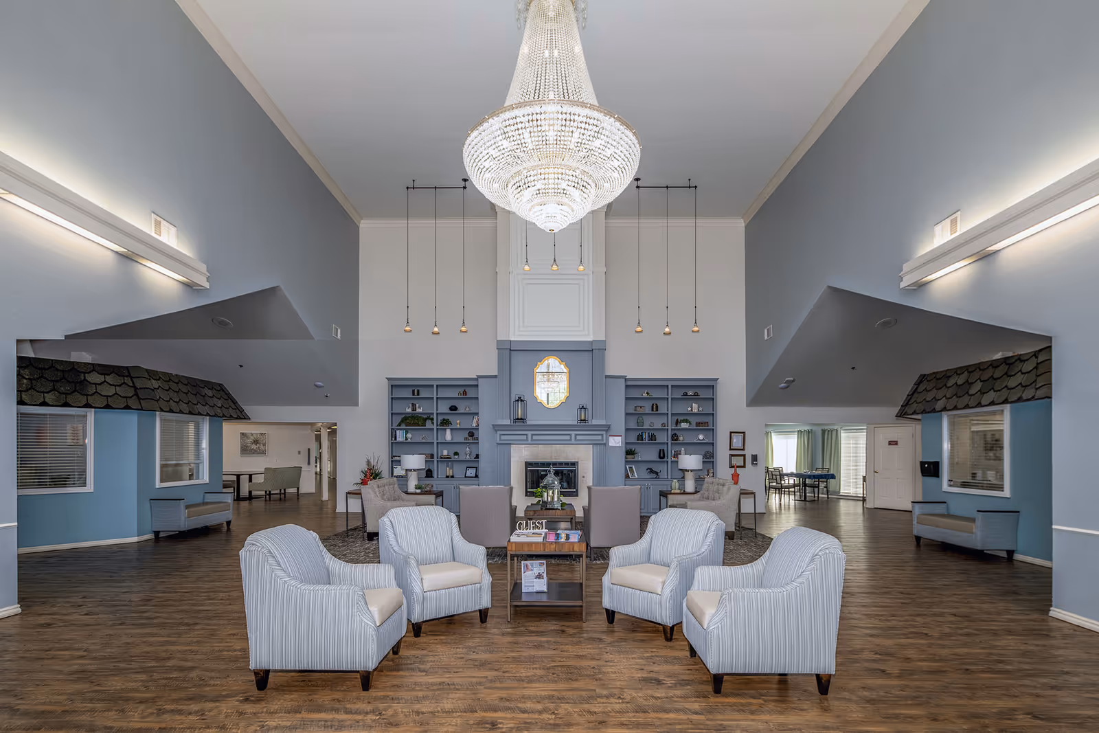 Spacious senior living facility common area with a large chandelier hanging from a high ceiling. The room features a seating arrangement with four striped armchairs around a small wooden table, additional chairs and sofas near a fireplace with built-in shelves on either side, and wood flooring throughout. The walls are painted light blue and white, and there are pendant lights hanging above the fireplace.