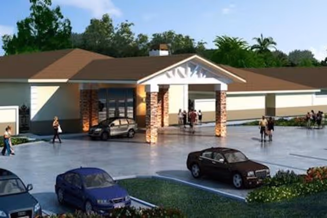 Exterior view of a single-story building with a covered entrance supported by brick columns. Several people are walking near the entrance and cars are parked in the parking lot in front of the building. Trees and greenery surround the area under a clear blue sky.