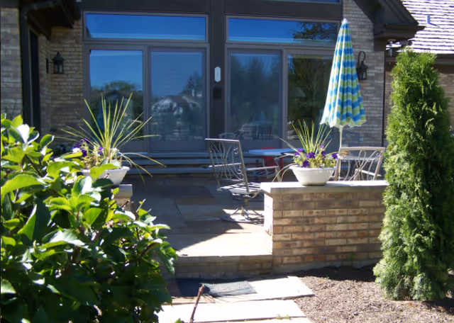 Outdoor patio area with a stone floor, metal chairs, a table with a blue and green striped umbrella, potted plants on brick pillars, and greenery surrounding the space in front of a building with large glass doors and windows.