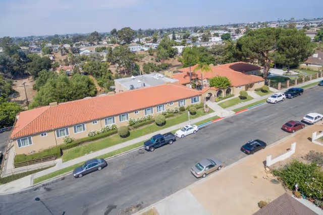 Aerial view of Arroyo Vista Nursing Center showing a single-story building with a red tile roof, surrounded by greenery and parked cars along the street in front. The building has multiple windows and an arched entrance area.
