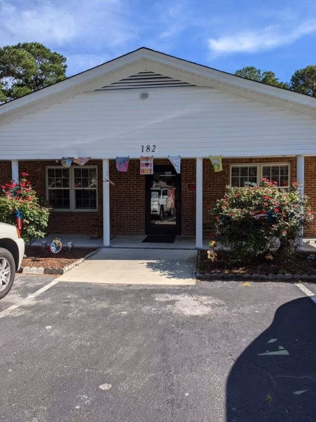 Front exterior view of a single-story brick building with white siding and a triangular roof peak. The building has a black door with the number 182 above it, flanked by two windows. There are colorful decorations hanging above the entrance and bushes with flowers on either side of the walkway leading to the door. Two parked vehicles are partially visible on either side of the image under a clear blue sky.