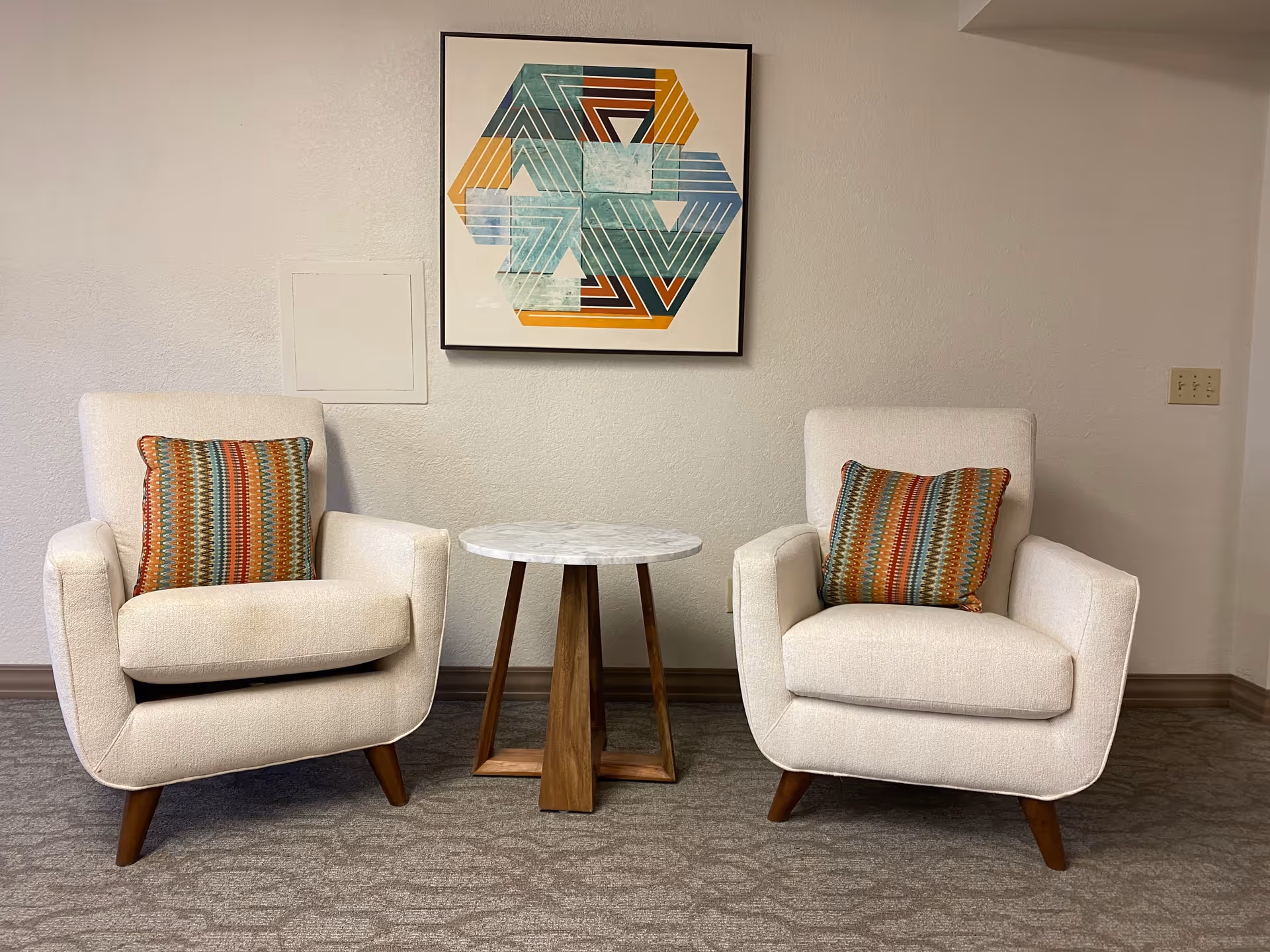 Two white upholstered armchairs with colorful patterned cushions are positioned on either side of a small round wooden table with a white marble top. Above the table hangs a framed geometric artwork featuring teal, orange, and brown colors. The setting is indoors with a neutral wall and carpeted floor.