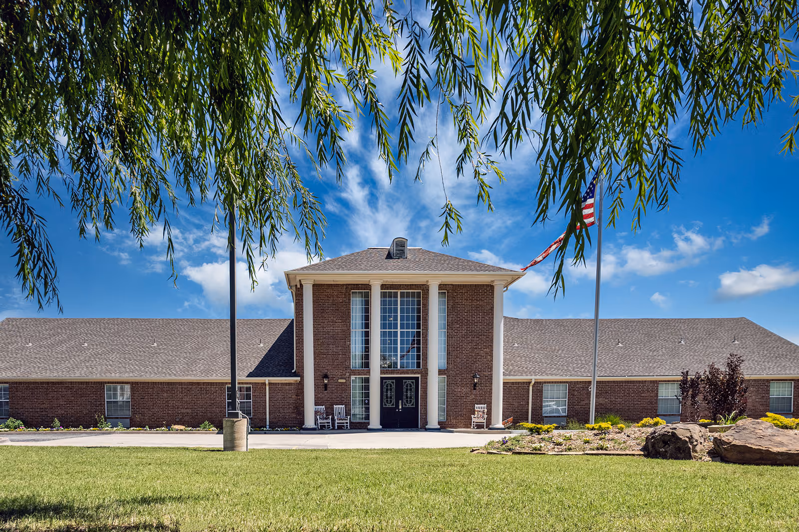 Front exterior view of a senior living facility named Sand Sage of The Highlands Senior Living, featuring a brick building with tall white columns, large windows, two rocking chairs near the entrance, an American flag on a flagpole, and green lawn with trees framing the top of the image under a blue sky with scattered clouds.