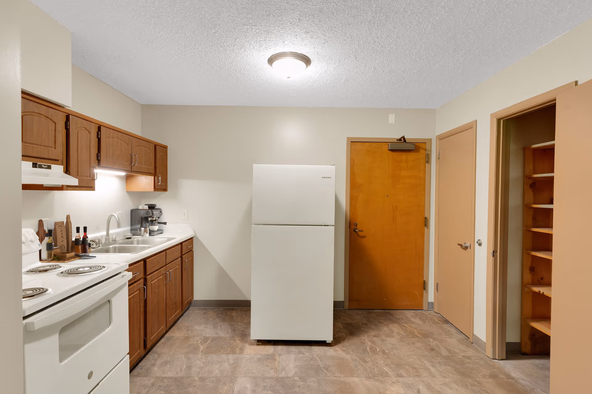 A kitchen with wooden cabinets, a white electric stove, a double sink, a white refrigerator, and a coffee maker. There are two closed doors and an open pantry with wooden shelves on the right side. The floor is tiled and the walls are painted beige.