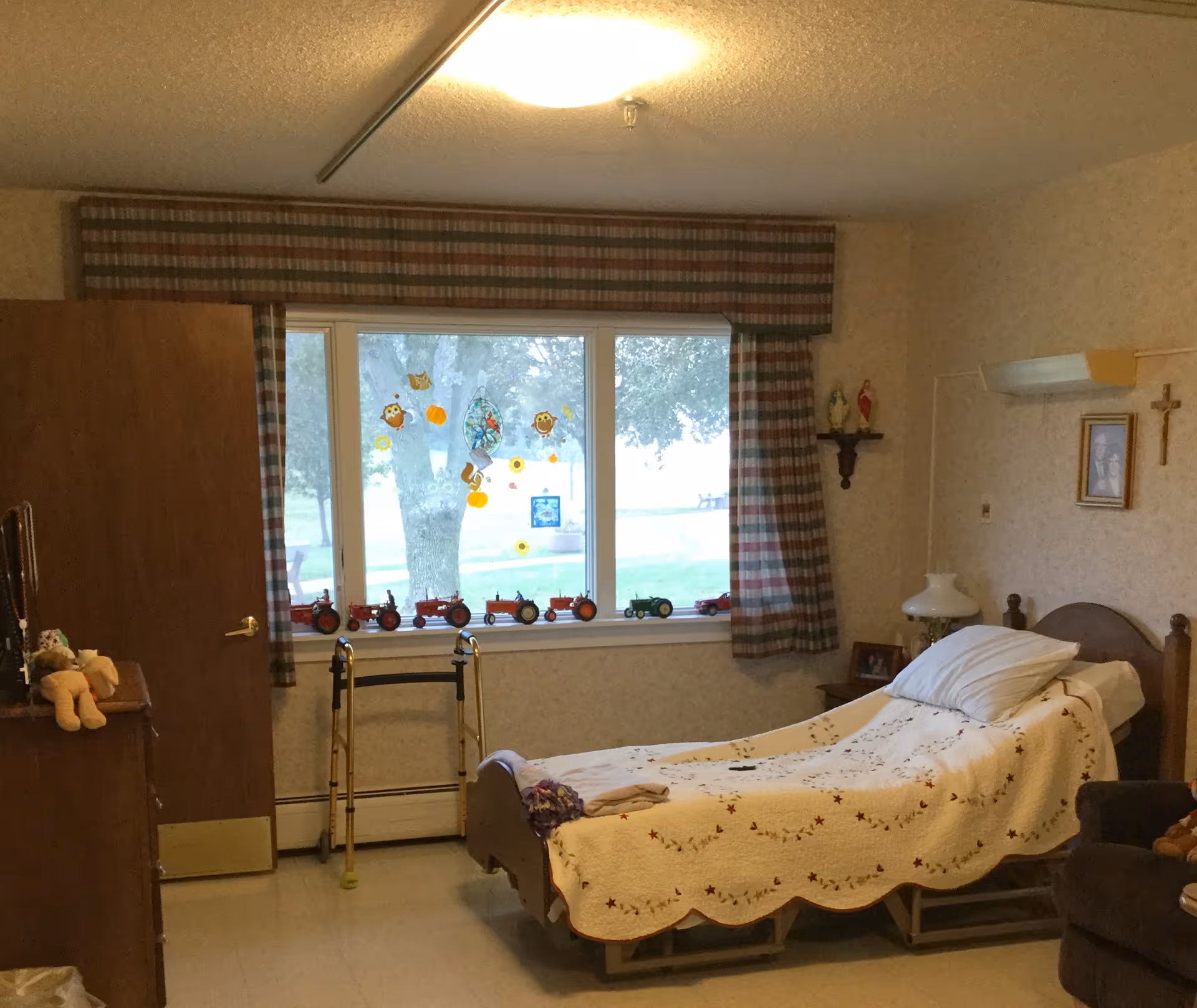A small senior bedroom with a single bed covered by a quilt, a walker by a large window decorated with toy tractors and plaid curtains, and a dresser.