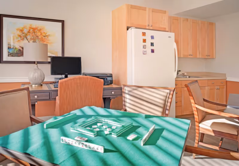 A well-lit room featuring a table with a green felt surface set up for a game of Mahjong, surrounded by wooden chairs. In the background, there is a small kitchenette with wooden cabinets, a white refrigerator, and a desk with a computer monitor, printer, and a lamp. A framed artwork hangs on the wall above the desk.