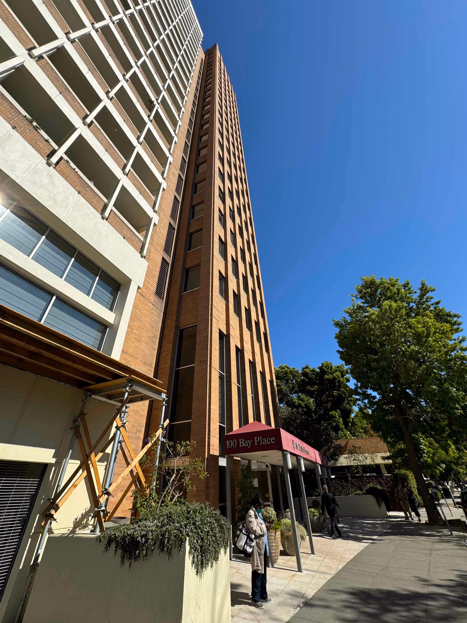 Tall brick building with multiple windows under a clear blue sky. A red canopy at the entrance reads '100 Bay Place'. A person wearing a mask and carrying a bag stands near the entrance, with trees and sidewalk visible nearby.