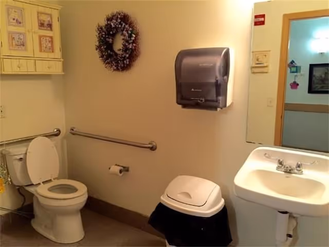 A bathroom with a toilet, a wall-mounted paper towel dispenser, a small sink with a faucet, a trash can with a lid, a handrail along the wall, a decorative wreath hanging on the wall, and a mirror reflecting part of an adjacent room.