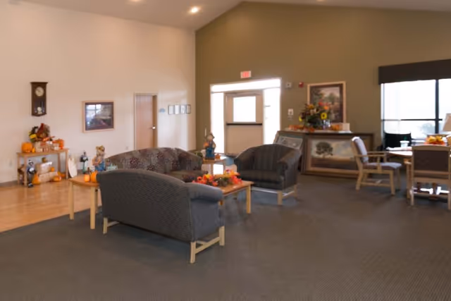 A cozy common area in a senior living facility with a seating arrangement including a patterned sofa and two armchairs around a wooden coffee table decorated with autumn-themed items. The room has a high ceiling, a wall clock, framed pictures, and a side table with seasonal decorations. There is a door with a window letting in natural light and a dining area with tables and chairs in the background.