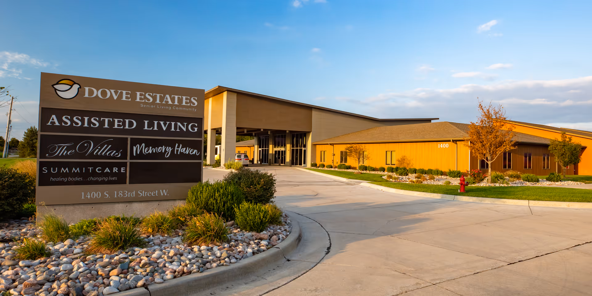 Exterior view of Dove Estates Senior Living Community building with a large sign in the foreground displaying the facility's name, services including Assisted Living, The Villas, Memory Haven, and SummitCare, along with the address 1400 S. 183rd Street W. The building is surrounded by landscaped greenery and a paved driveway under a partly cloudy sky.