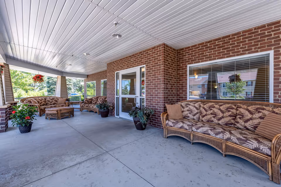 Covered outdoor patio area with wicker furniture including sofas and a coffee table, potted plants, and a brick wall with windows and a door leading inside. Trees and greenery are visible in the background.
