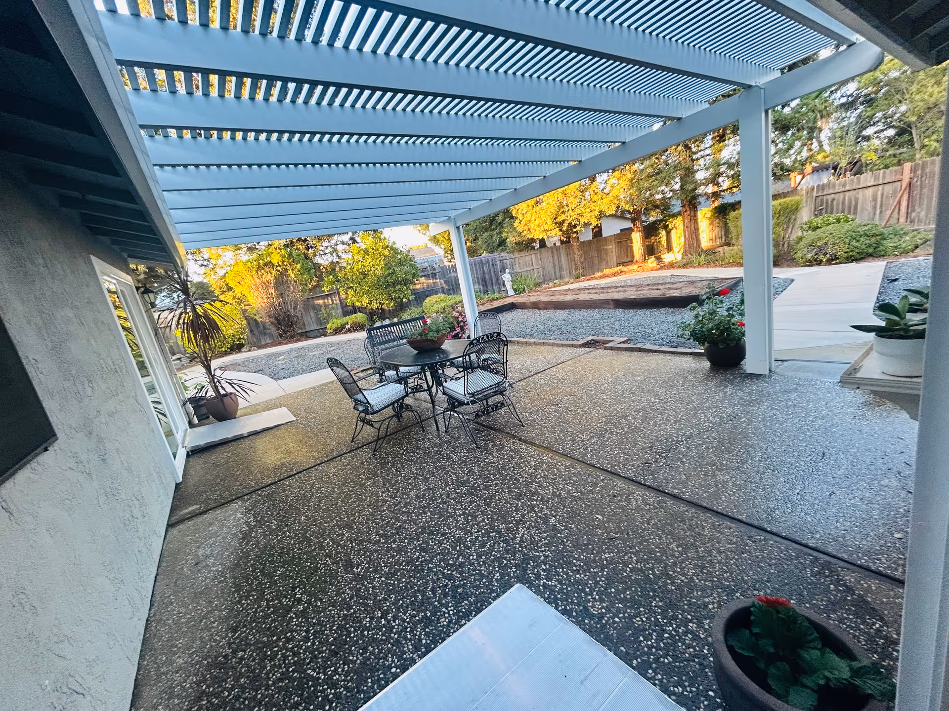 Outdoor patio area with a white pergola overhead, a round metal table with four chairs, potted plants, and a landscaped garden with trees and bushes in the background.