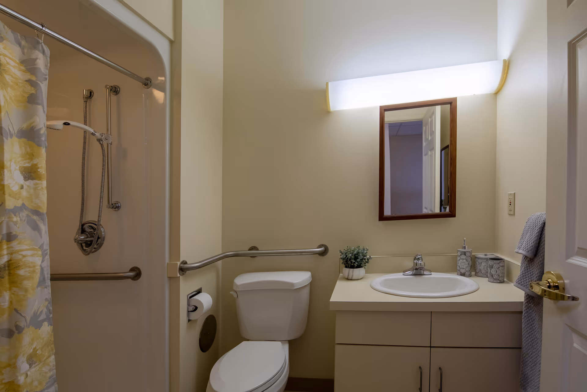 A bathroom with a toilet, a sink with a countertop, a mirror above the sink, and a shower with a yellow and gray floral shower curtain. The bathroom has grab bars near the toilet and in the shower area, a towel hanging on the wall, and a small potted plant on the countertop.