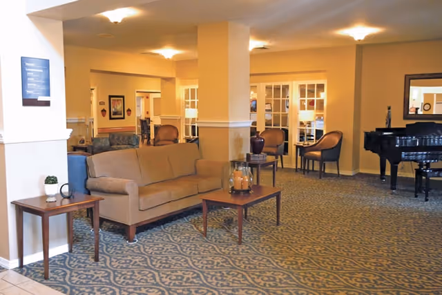 Lobby-style seating area with a sofa, armchairs, coffee tables, and a grand piano in a well-lit common room.