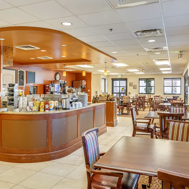 A spacious dining area with multiple wooden tables and chairs arranged neatly. The room features a counter with various condiments, coffee machines, and cups. The ceiling has recessed lighting and hanging pendant lights. Large windows allow natural light to brighten the space.