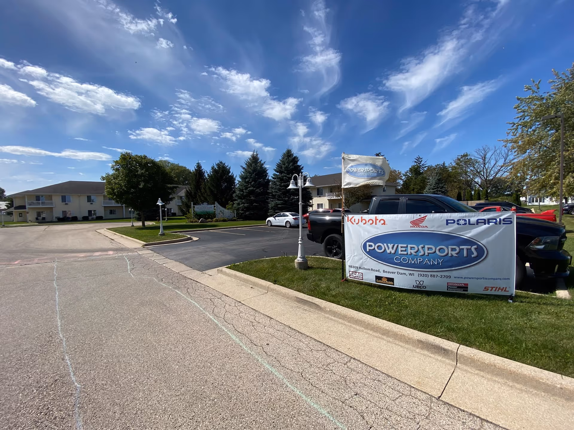 Exterior view of a senior living facility with a parking lot, a black pickup truck, and a large banner advertising Powersports Company. The sky is blue with scattered clouds, and there are trees and a two-story building in the background.
