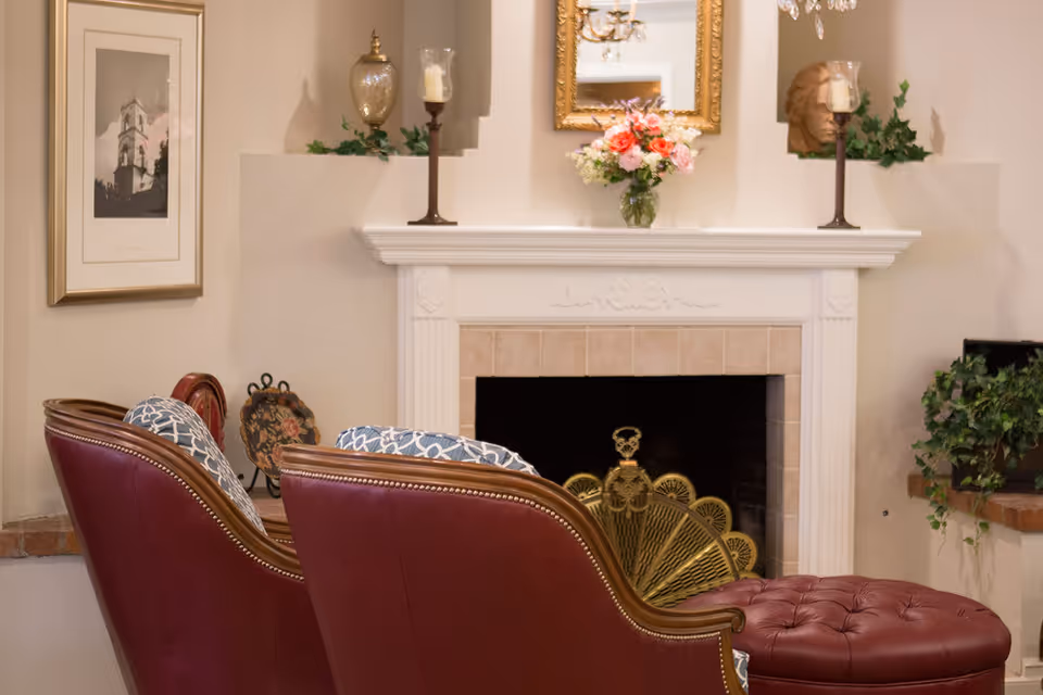 Cozy living room with a white fireplace mantel, decorative items, two red upholstered chairs and a tufted ottoman.