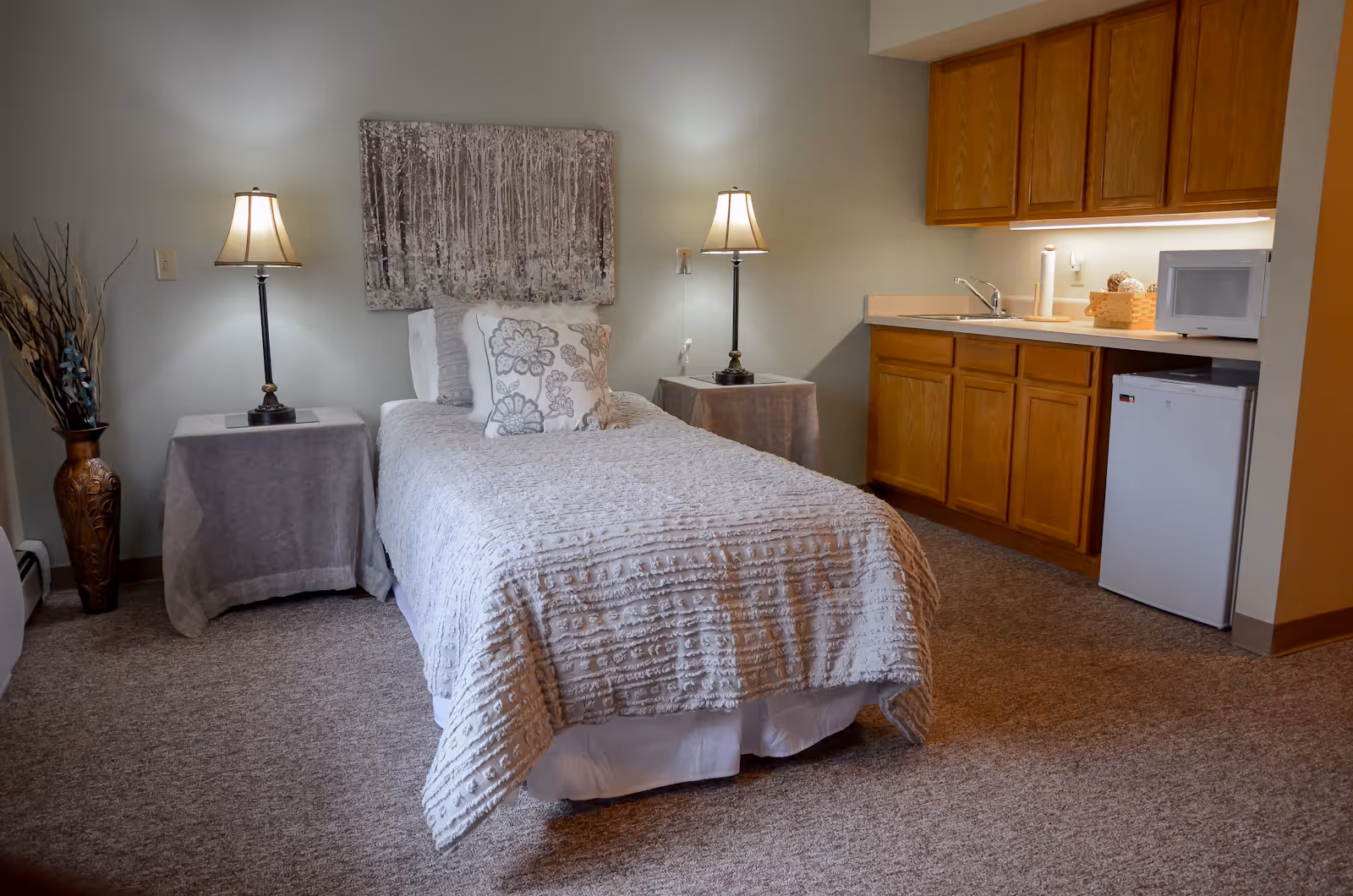 A cozy senior living bedroom with bed, nightstands, lamps, and kitchenette.