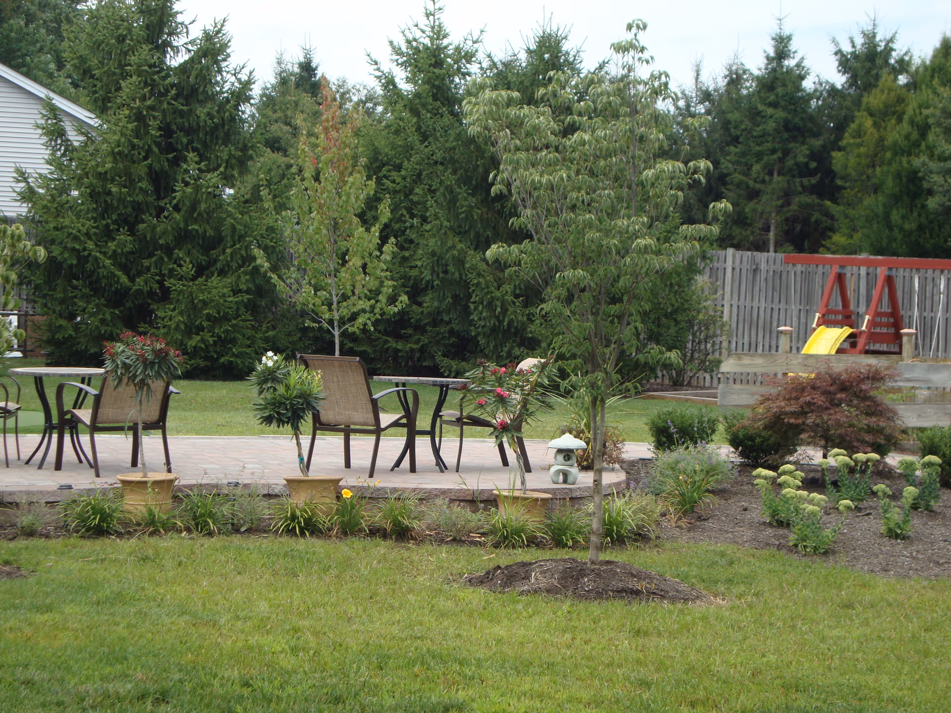 Outdoor garden area with green grass, small trees, and various plants. There are patio tables and chairs on a paved area, and a wooden swing set with a yellow slide is visible in the background. The area is surrounded by tall evergreen trees and a wooden fence.