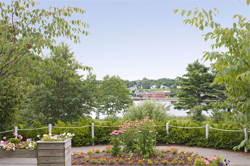 A garden area with colorful flowers and green shrubs, bordered by a rope fence. In the background, there are trees and a body of water with buildings on the far shore under a clear sky.