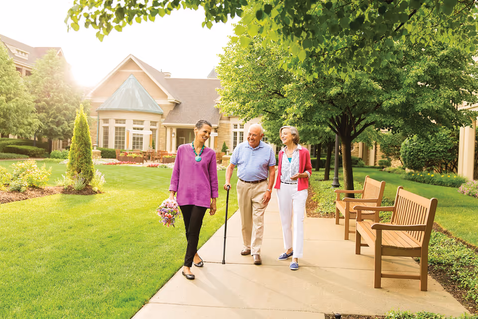 Three elderly people walking and talking on a paved pathway in a lush garden area with green grass, trees, and benches. A large building with a peaked roof and multiple windows is in the background, and the sun is shining brightly.