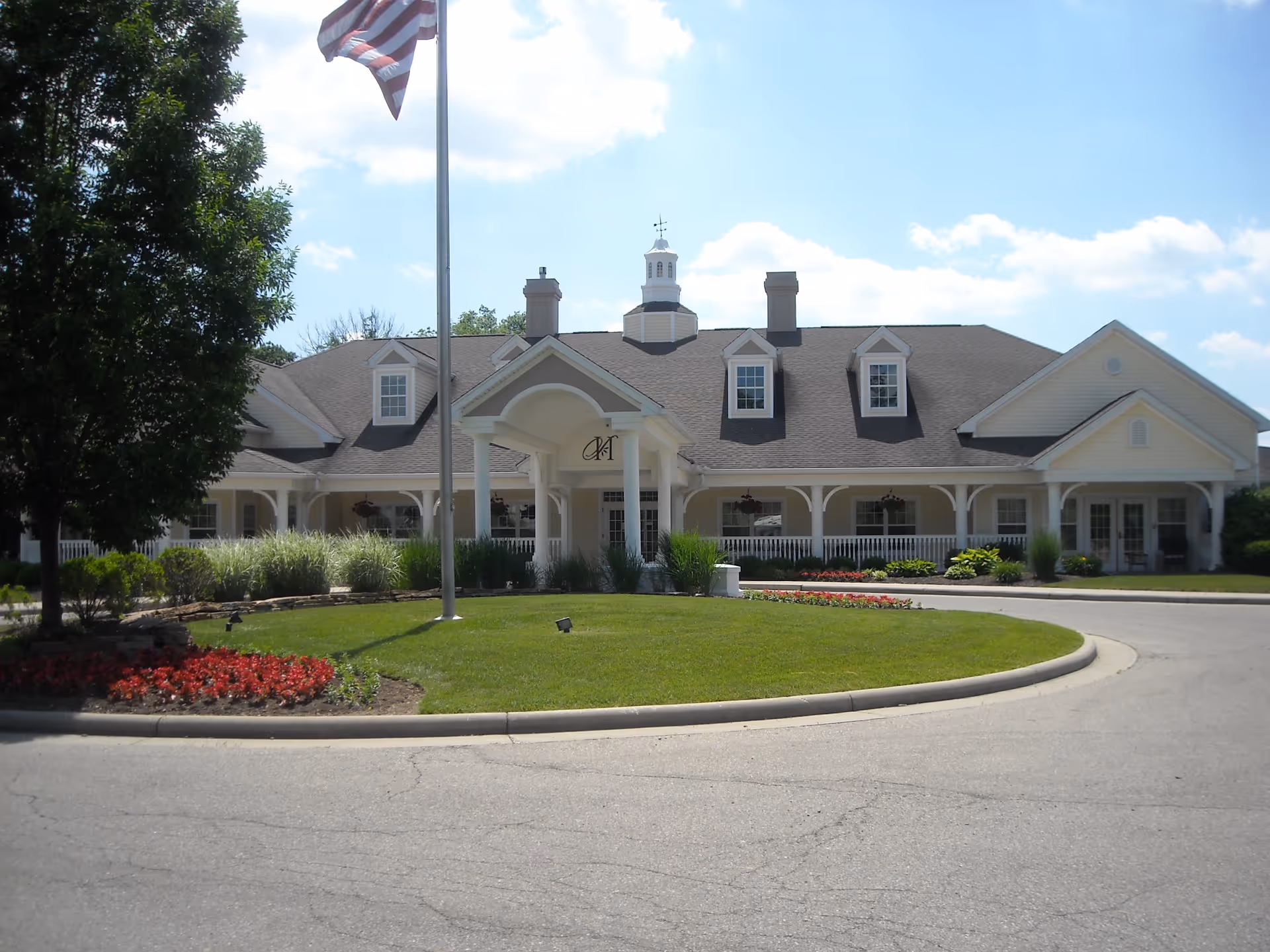 Front exterior view of a single-story building with a pitched roof, dormer windows, and a covered entrance supported by columns. There is a flagpole with an American flag in front of the building, surrounded by a well-maintained lawn and flower beds. The sky is partly cloudy.
