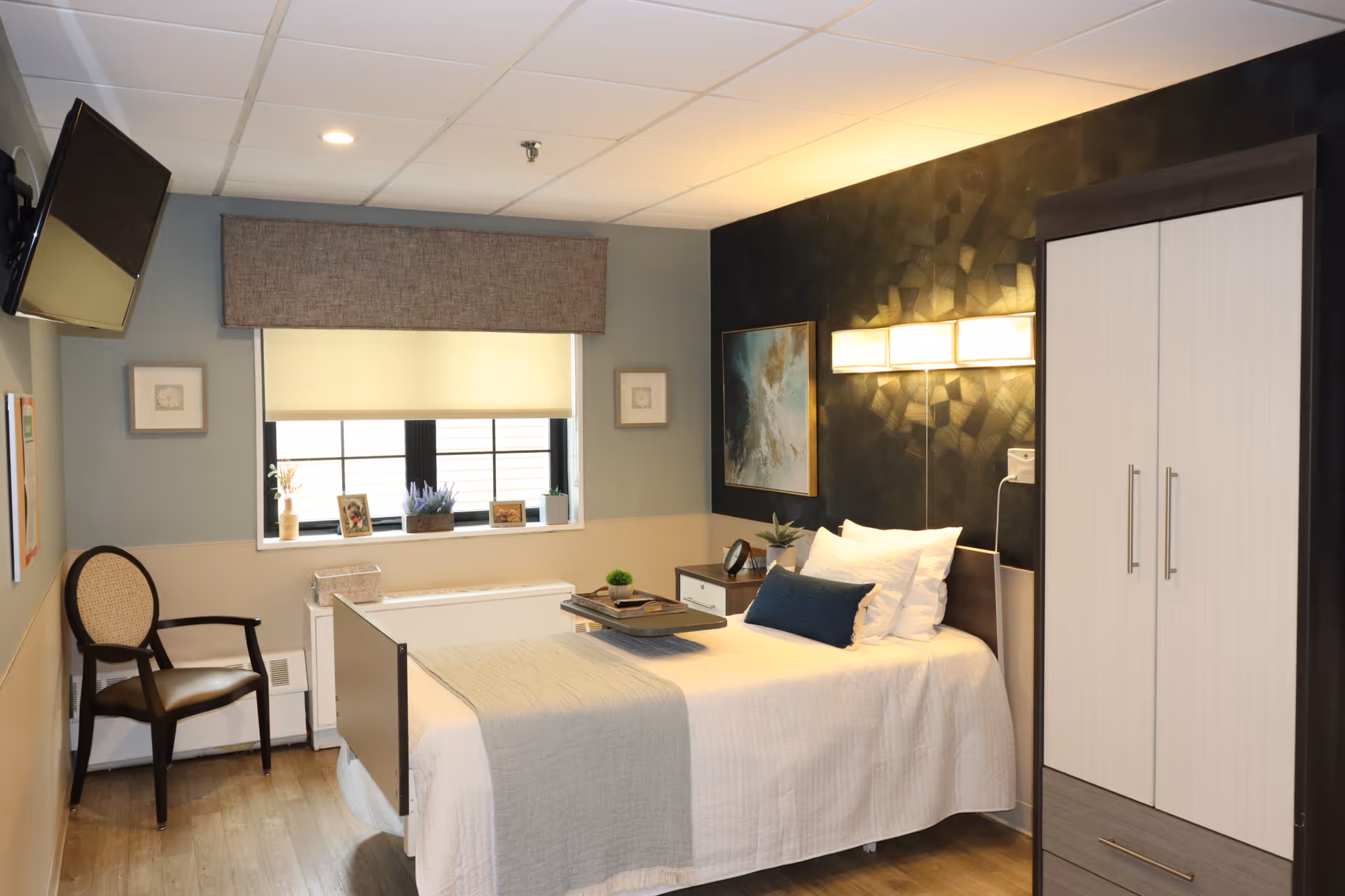 A neatly arranged bedroom in a senior living facility with a single bed made up with white and beige bedding, a blue accent pillow, a bedside table with a clock and plant, a wardrobe, a chair, and a window with a beige roller shade and valance. The walls are decorated with framed artwork and the room has wood flooring.