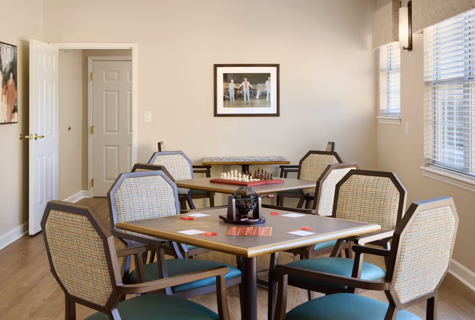 A bright communal activity room with multiple tables and upholstered chairs, a chess set and cards on the tables, and windows letting in natural light.