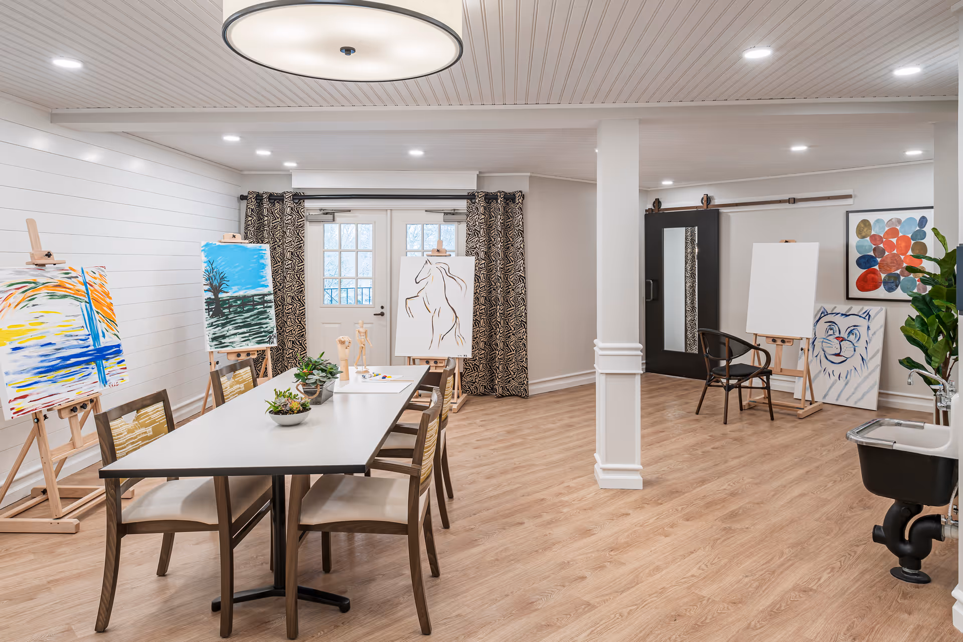 A bright and spacious art room with wooden flooring and white walls. The room features a long table with chairs around it, several easels displaying colorful paintings, a black chair near a blank canvas, a water fountain, and decorative plants. Large windows with patterned curtains allow natural light to enter the room.