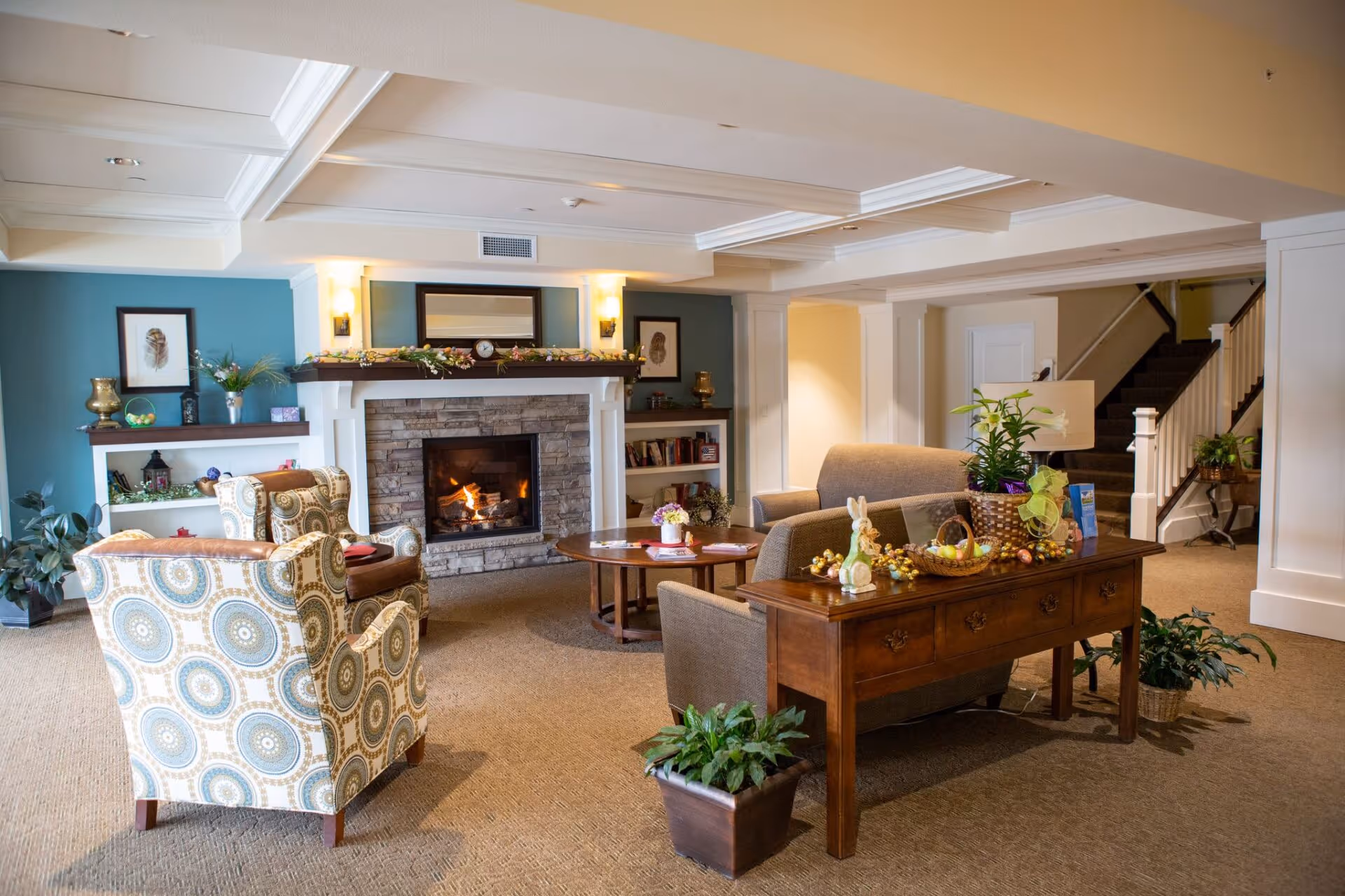 A cozy living room area in AVIVA River Bend featuring a lit stone fireplace with a wooden mantle decorated with garlands. The room has two patterned armchairs and a gray sofa around a wooden coffee table. There are plants and decorative items on a wooden console table behind the sofa. The walls are painted in soft colors with built-in shelves holding books and decorations. A staircase is visible in the background.